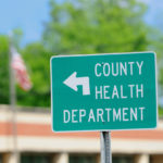 Photograph of a green county health department directional street sign with building and street light in background